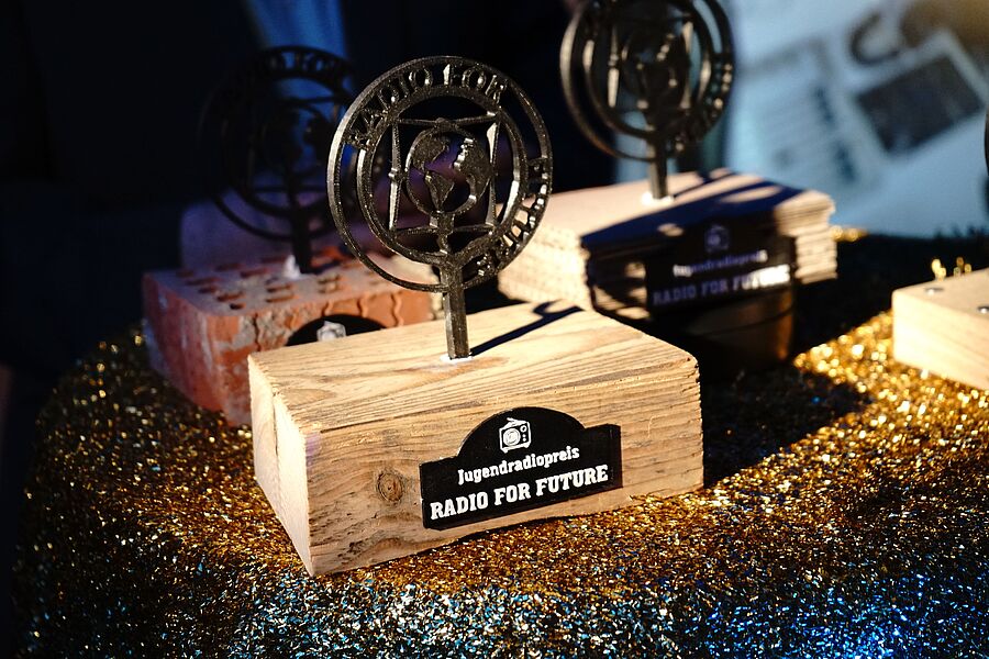 Preise aus Holz mit metallischen Emblemen stehen auf einem glitzernden Tisch, beschriftet mit „Radio for Future“.