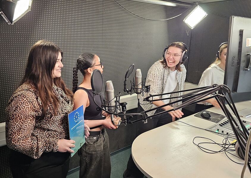 Drei Frauen stehen in einem Tonstudio, lächeln und sprechen in Mikrofone, während eine von ihnen ein Dokument hält.